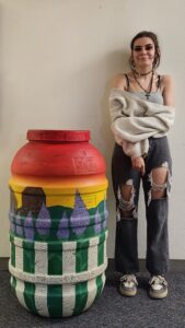 Cadence Larson with her painted rain barrel, which depicts a sunset and homestead, with purple flowers and a white fence in the foreground