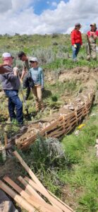 Using a skidsteer to create a slope on the downhill side of a rock core wicker weir, to control the grade below a log and fabric structure